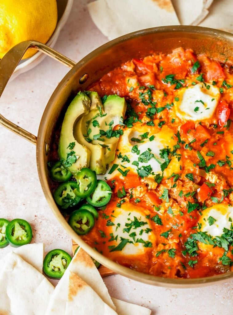 A pan of shakshuka with poached eggs in a rich tomato sauce, garnished with fresh parsley, sliced avocado, and jalapeños