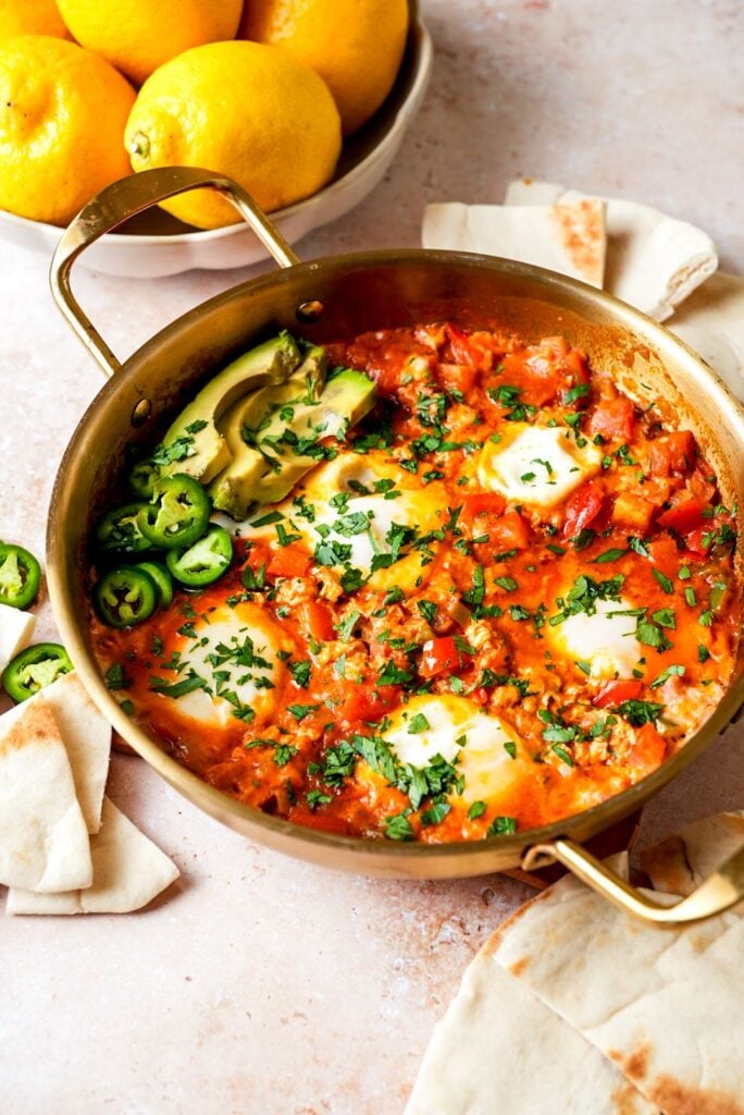 A pan of freshly made shakshuka with poached eggs in a rich tomato sauce, garnished with fresh parsley, sliced avocado, and jalapeños, served with warm pita bread on the side