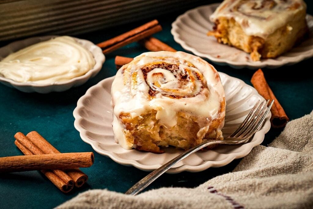 A soft, fluffy cinnamon roll topped with creamy icing on a white scalloped plate, surrounded by cinnamon sticks and a dish of frosting
