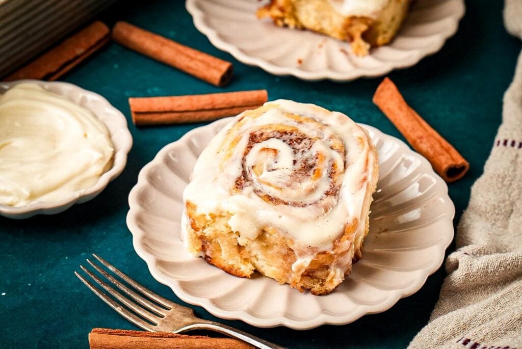 A soft buttery cinnamon roll topped with creamy icing on a white scalloped plate, surrounded by cinnamon sticks and a dish of frosting