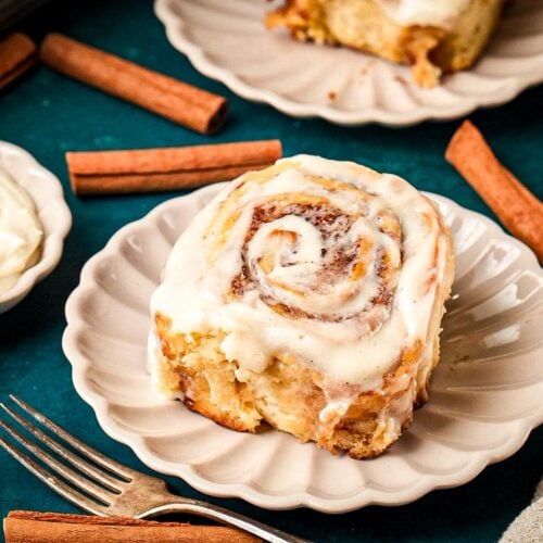 A soft buttery cinnamon roll topped with creamy icing on a white scalloped plate, surrounded by cinnamon sticks and a dish of frosting