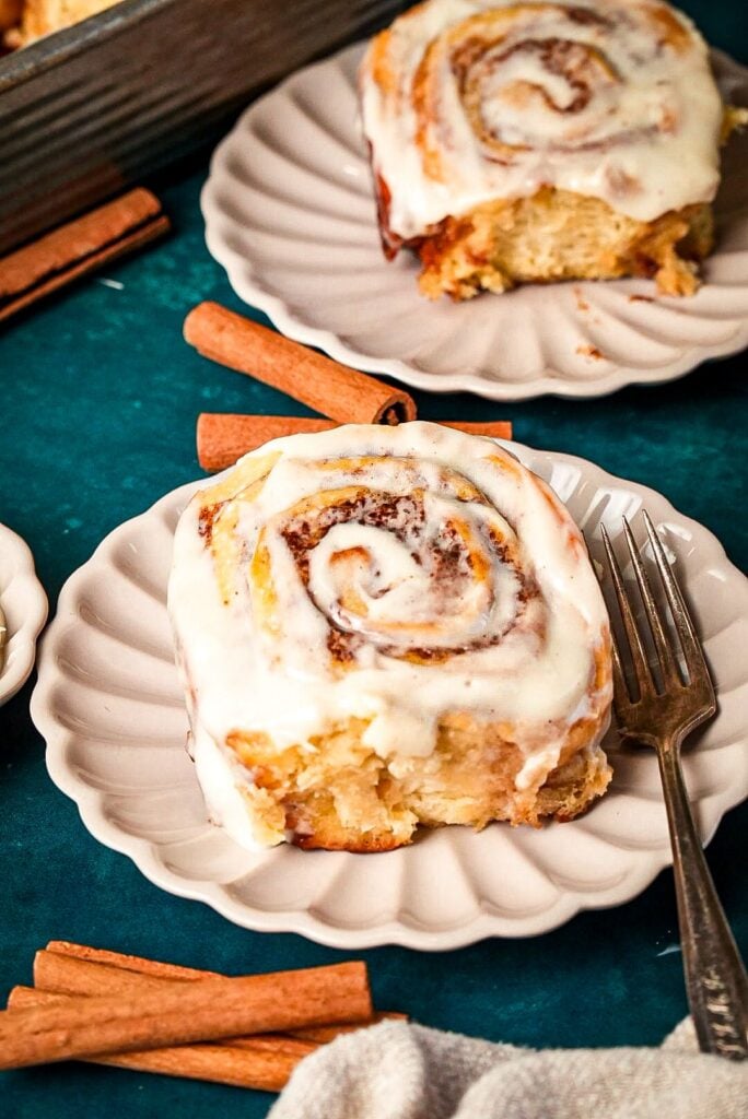 A shot of two serving plates for soft, fluffy cinnamon rolls topped with creamy icing, surrounded by cinnamon sticks