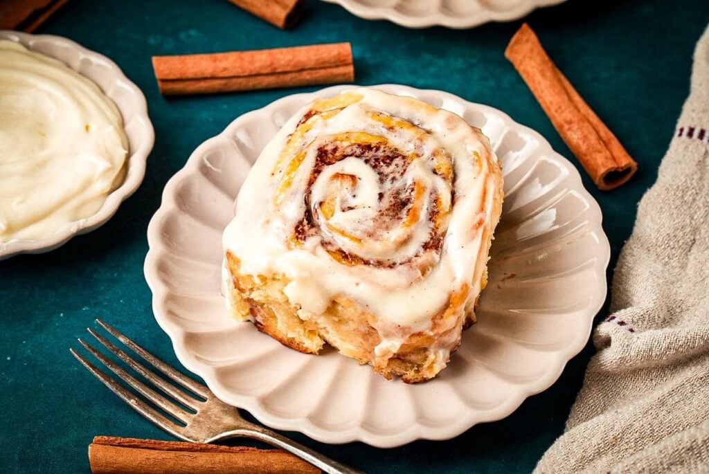 A fluffy cinnamon roll topped with creamy icing on a white plate, surrounded by cinnamon sticks and a dish of frosting