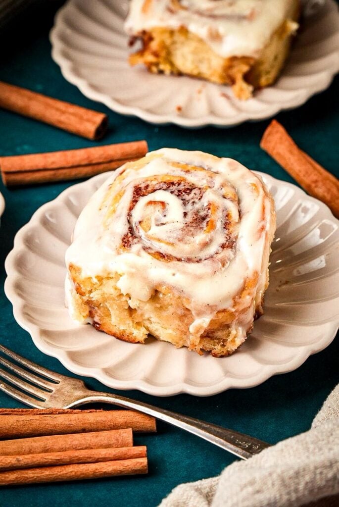 A shot of a serving plate for soft, fluffy cinnamon rolls topped with creamy icing, surrounded by cinnamon sticks next to another plate of cinnamon rolls