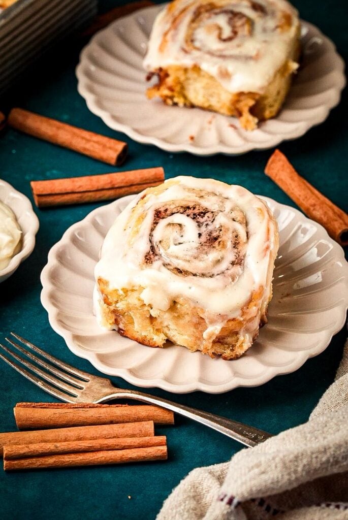 A shot of two serving plates for soft, fluffy homemade cinnamon rolls topped with creamy icing, surrounded by cinnamon sticks